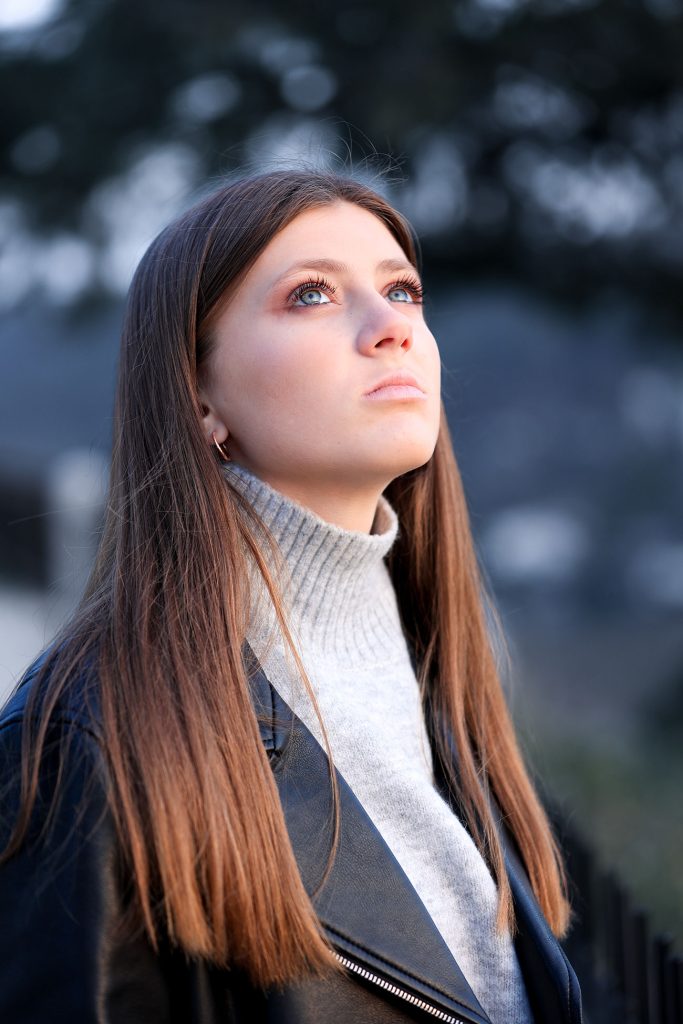 Jeune femme aux cheveux longs regardant vers le ciel avec un regard intense, vêtue d'un blouson noir et d'un col roulé gris clair, portrait en extérieur avec un fond flou bleuté