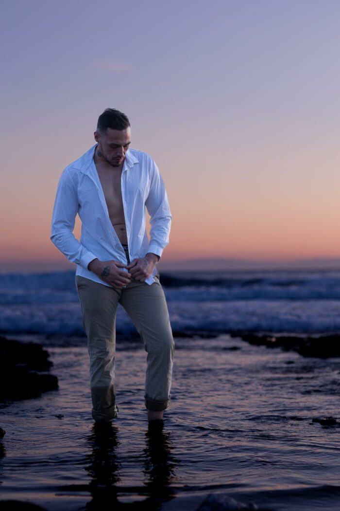 Homme en pantalon beige et chemise blanche ouverte sur ses abdominaux, pieds dans l'eau, photo prise pendant le golden hour à la plage de Carro à Martigues